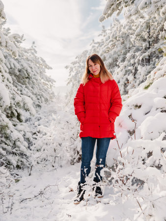 Young woman in red jacket among snowy trees in winter forest. Traveler woman with snow trees in...