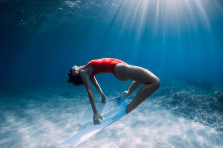 Woman freediver in red swimsuit with white fins glides underwater over sand in tropical ocean. Sporty girl dive underwater.の写真素材