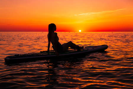Slim woman on stand up paddle board at quiet sea with bright sunset or sunrise. Woman on sup board and sunset with reflectionの写真素材