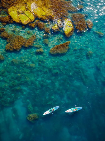 May 28, 2021. Anapa, Russia. Couple on stand up paddle board at blue sea with rocks. People walking on Red paddle sup board in transparent sea. Aerial viewのeditorial素材