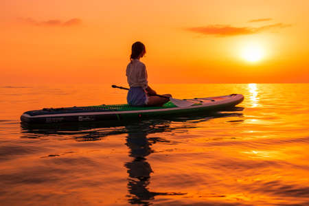 June 21, 2021. Anapa, Russia. Sporty girl on stand up paddle board at quiet sea with sunset or sunrise. Woman on Red Paddle sup board and bright sunsetのeditorial素材