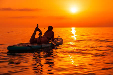 June 21, 2021. Anapa, Russia. Sporty girl on stand up paddle board at quiet sea with sunset or sunrise. Woman on Red Paddle sup board and bright sunsetのeditorial素材