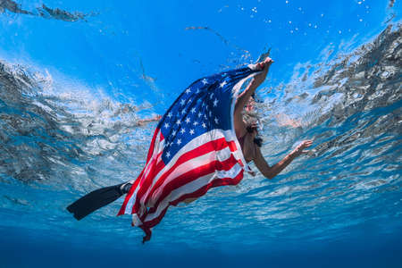 Woman with fins swimming in tropical ocean with United States flag. Independence day concept.