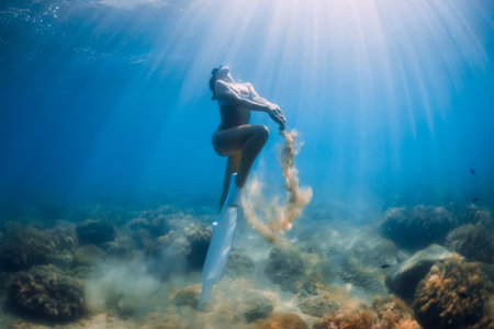 Freediver underwater glides with white fins over sandy bottom. Attractive woman free diver in blue ocean with sun raysの写真素材