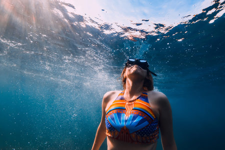 August 07, 2021. Varna, Bulgaria. Sporty woman with freediving fins posing underwater in ocean with sunlight. Young girl in colorful bikini glides underwater.の写真素材