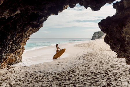 Surf girl with surfboard posing on ocean beach. Woman with surf board and cave rocks in Baliの写真素材