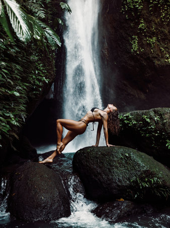 Gorgeous slim woman in bikini posing near waterfall. Traveler girl posing on waterfallの写真素材