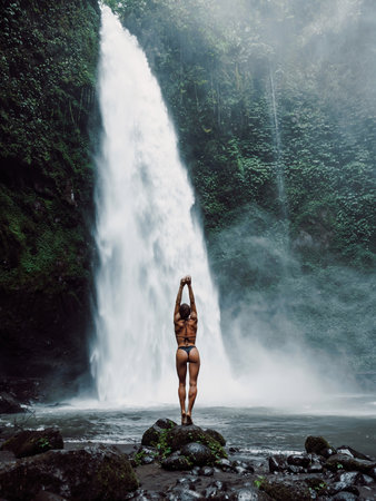 Slim woman in bikini posing near waterfall in tropical Bali. Traveler girl on powerfull waterfallの写真素材