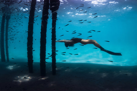 Woman freediver underwater with fishes in blue sea. Female swims under the pier with pier basesの写真素材