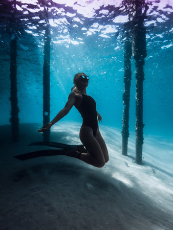 Freediver woman posing underwater under the pier in blue ocean. Female swims with fins under the pierの写真素材