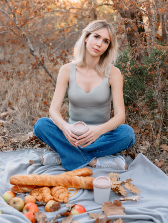 Attractive blonde woman sitting on a plaid with drink and bakery on a picnic in forest with warm sunshineの写真素材