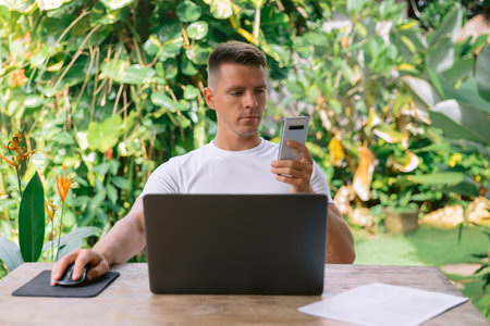 Serious man holding a smartphone and working on a laptop in an open plan office.