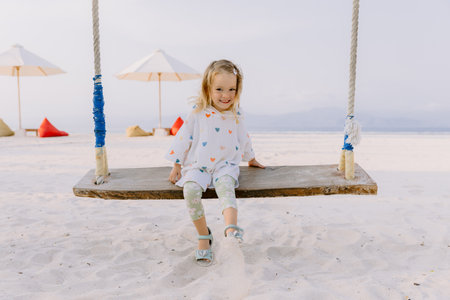Little child girl plays and swinging on swings on tropical beach, enjoy childhoodの写真素材