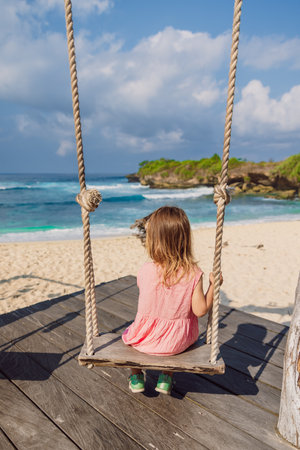 Little child girl plays and swinging on swings on tropical beach with ocean, enjoy childhoodの写真素材