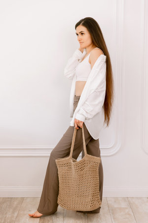 Elegant woman with long hair posing barefoot, carrying a stylish woven tote bag, showcasing casual chic fashion indoorsの写真素材