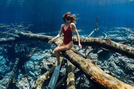 Woman free diver sitting underwater on sunken log in crystal blue water. Freediving in fresh water lake in Indonesiaの写真素材