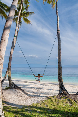 Woman on a tropical beach swing, enjoying the ocean view and freedom under palm trees, a perfect island escape.の写真素材