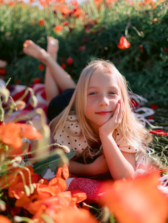 Blonde child girl lying on a plaid in a blooming poppy field, sunny summer dayの写真素材