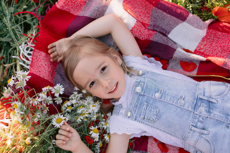 Smiling little girl lying on a picnic blanket among wildflowers, enjoying a sunny summer day in nature.の写真素材