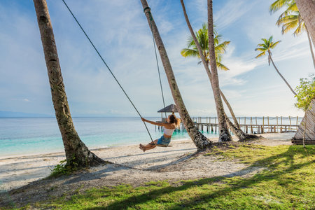 Woman swinging on a tropical beach, enjoying freedom and sunshine under palm trees, paradise vibes.の写真素材