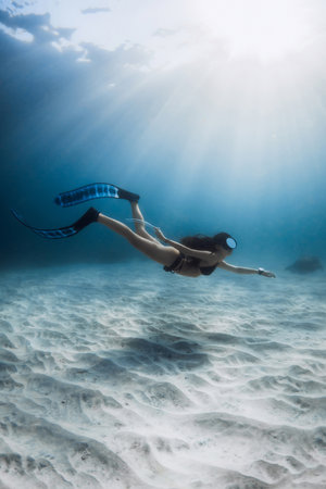 Woman underwater with diving fins swims in blue sea. Freediving over white sand sea bottom, activity water sport.の写真素材