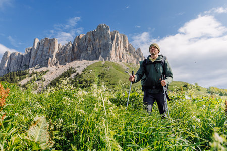 Male hiker with trekking poles stands in green meadow with rocky mountains under blue sky.の写真素材