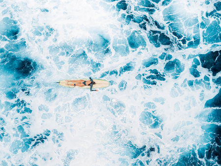 Woman swims on surfboard in blue ocean with foam waves. Drone view of woman during surfingの写真素材