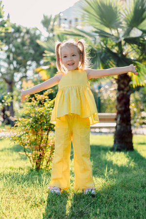 Happy little girl in yellow dress enjoying summer in sunny park, arms wide open, smiling brightly.の写真素材
