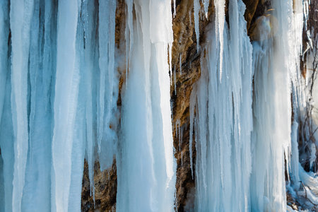 Frozen waterfall with long icicles on a rocky cliff in winter sunlight.の写真素材