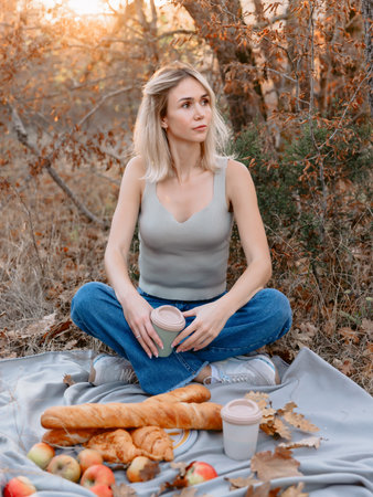 Attractive caucasian woman holding cup of coffee and sitting on a plaid with bakery on a picnic with warm sunshineの写真素材