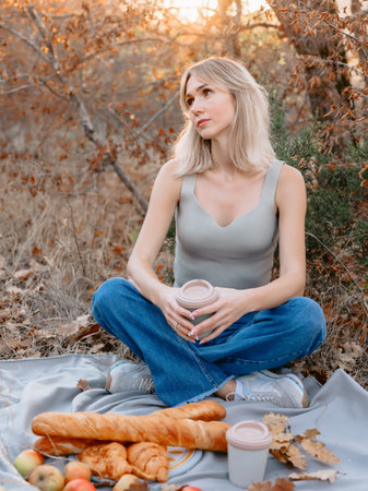 Caucasian woman holding cup of coffee and sitting on a plaid with bakery on a picnic with warm sunshineの写真素材