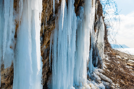 Stunning long blue icicles hanging from a rocky cliff face by a frozen lake shore in bright winter sunlight.の写真素材