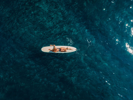 January 25, 2026. Bali, Indonesia. Aerial view of surfer girl floating on longboard in blue ocean....