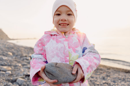 Smiling child in warm clothes holds a stone on a rocky beach at sunsetの写真素材