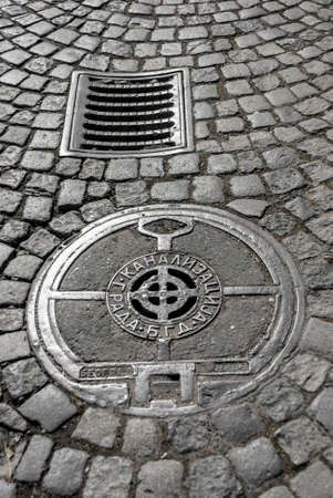 Cast iron storm drain and sewer manhole cover on cobblestone street. Content cyrillic caption: "City Sewage B.G.D.". "B.G.D." is stands for Belgrade, Serbiaの写真素材