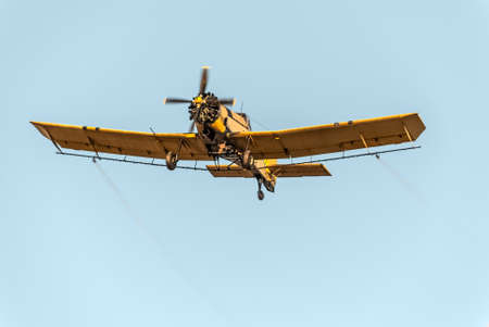 Camera catches a yellow crop duster in flight, while spraying mosquitoes. Low angle view over clear blue sky. All inscriptions (company names and technical informations) have been carefully removed.の写真素材