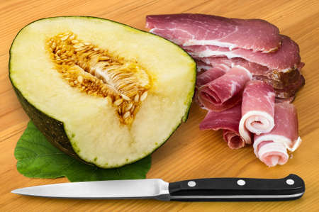 Salty prosciutto, sweet melon with leaf and kitchen knife on wooden background in slightly rustic mediterranean scene.の写真素材