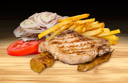 Grilled pork chop with French fries, sliced onion, and tomato on a wooden background. This is a high-resolution, dark scene.の写真素材