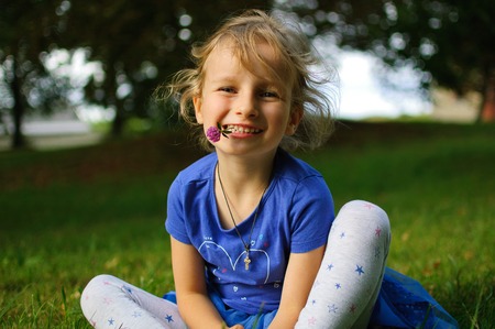 Lovely curly girl with light brown hair is sitting on the grass. She holds a purple clover flower in...