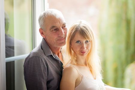 Portrait of Senior Man Hugging His Young Wife on Windows Background Indoorsの写真素材