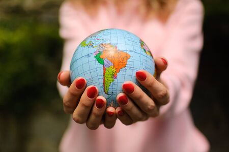 The girl in a pink sweater with red manicure holds a small globe with geographical names in Ukrainian cyrillic letters on it. South America is visible. Ecological problems concept.の写真素材