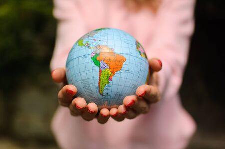 The girl in a pink sweater with red manicure holds a small globe with geographical names in...