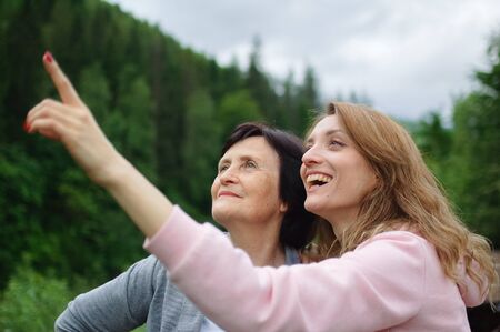 Happy senior mother and adult daughter are travelling and posing together over landscape of forest and mountains, young girl is pointing at something. Concept of travelling with familyの写真素材