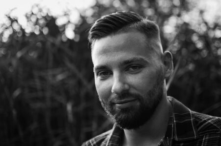Black and white portrait of young bearded man looking at the camera outdoors in the park on trees backgroundの写真素材