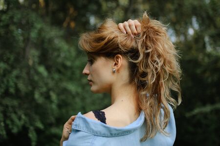 Back view of blonde adult woman wearing blue casual jeans blouse posing in the park on green trees background. Hipster style concept.の写真素材