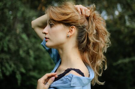 Side view portrait of sexy blonde girl with curly hair wearing blue casual jeans blouse and black lingerie posing in the park on green trees background. Hipster style concept.の写真素材