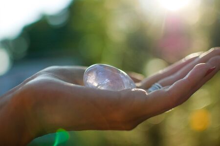 Female hand with transparent amethyst quartz yoni egg for vumfit, imbuilding or meditation. Shining crystal egg in hands on sky and sunrise background outdoors. Womens health concept.の写真素材