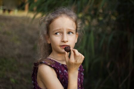 Beautiful little blond girl in violet dress with ponytail is eating a chocolate candy in the park durring a day, sugar addiction conceptの写真素材