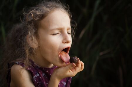 Beautiful little blond girl in violet dress with ponytail is eating a chocolate candy in the park durring a day, sugar addiction conceptの写真素材
