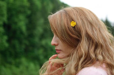 Outdoors portrait of young calm blonde woman in pink clothes with little yellow flower in her long...